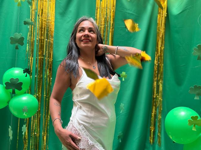 Laila Castillo, una mujer de curvas con cabello oscuro, lleva un vestido blanco de encaje, posando con confianza y con una mano en la cadera, verde. globos y tréboles en el fondo, flores amarillas en movimiento.