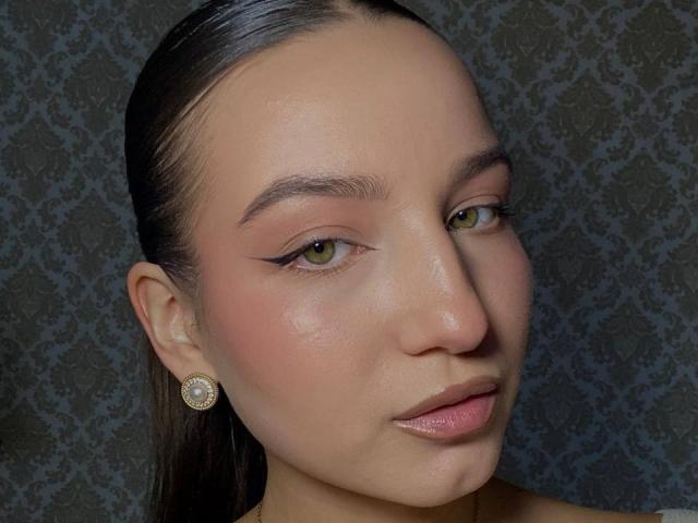 Close-up of MargertLewi's face with slicked-back hair, green eyes, pink blush, and glossy pink lips. She wears a round, jeweled earring on her left ear. Fucking sexy, intense gaze.