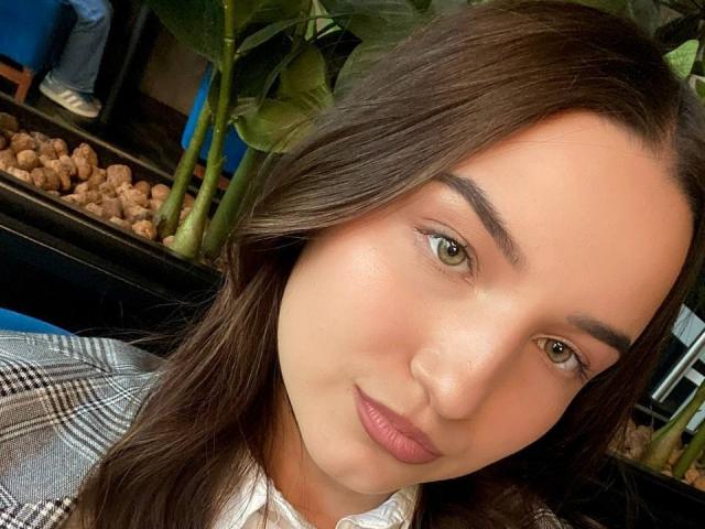 Close-up selfie van MargretLewi, met lang donker haar, groene ogen en roze lippen. Ze draagt een geruit blazer over een wit shirt, met stukken gemberwortel op de onscherpe achtergrond. Heel erg aantrekkelijk..