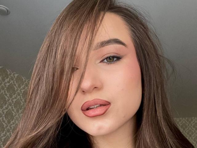 Nahaufnahme von MargretLewi, braunes Haar bedeckt ein Auge, grüne Augen, rosafarbene Lippen leicht geöffnet, wobei ein Hauch von Zunge sichtbar ist. Ihre Haut ist blass, ihr Haar ist glänzend, der Hintergrund ist grau gemustert..