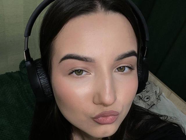 Close-up of MargertLewi with fair skin, green eyes, and dark brown hair, wearing black headphones, pouting with pink lips. Her expression exudes a slight sexual tension. Soft lighting highlights her face.