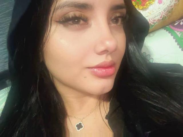 Close-up selfie van AmaliaDalton, een jonge vrouw met lang zwart haar, bruine ogen en roze lippenstift. Ze draagt een kleine bloemenketting en kijkt recht in de camera met een verleidelijke uitdrukking..