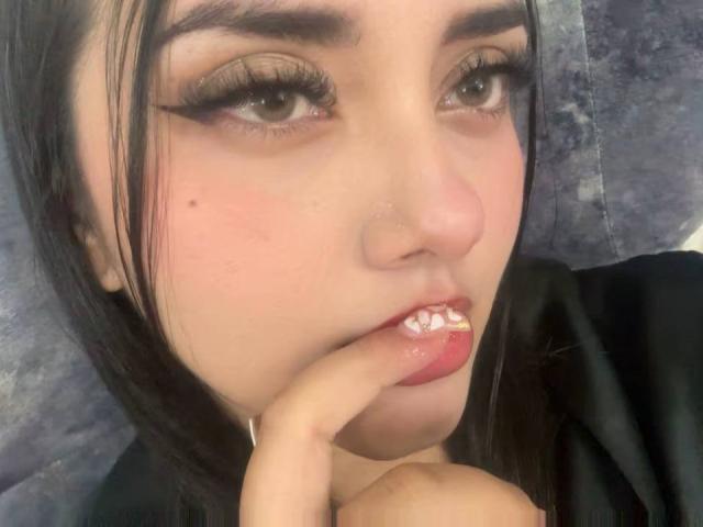 Fotografía de primer plano de AmaliaDalton, con piel pálida, cejas oscuras y cabello negro largo, mordiéndose los labios rosados y brillantes. dedo índice, mirando seductoramente a la cámara, contacto visual intenso.