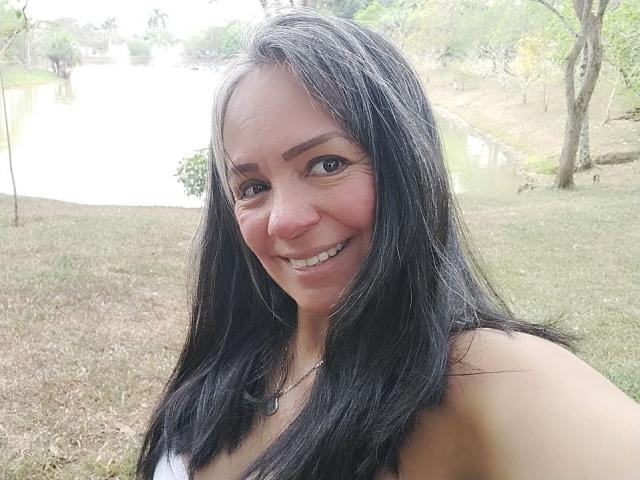 Fotografie von Laila Castillo mit langen, schwarzen Haaren, die lächelt, mit freiliegenden Schultern, im Freien in einem grasbewachsenen Park mit Bäumen. Helles Sonnenlicht, leichte Röte auf den Wangen, natürliches Licht betont ihr Gesicht..