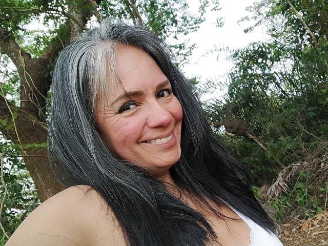 Close-up of LailaCastillo, a middle-aged woman with long black hair and gray highlights, smiling with a flirtatious look. She has tan skin and is wearing a white top. Outdoor setting with trees.