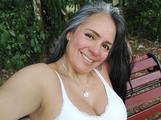 Foto van Laila Castillo met lang zwart haar, lachend, gekleed in een witte kanten bh, waarbij haar decolleté goed zichtbaar is. Ze zit op een rode bank in een bebost gebied, met natuurlijk licht. Heel aantrekkelijk..