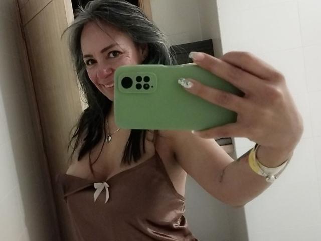 Laila Castillo prenant un selfie dans un miroir, tenant un téléphone vert avec une coque blanche. Elle porte un caraco en satin marron avec un nœud, qui dévoile sa poitrine. Cheveux noirs, souriant
.
