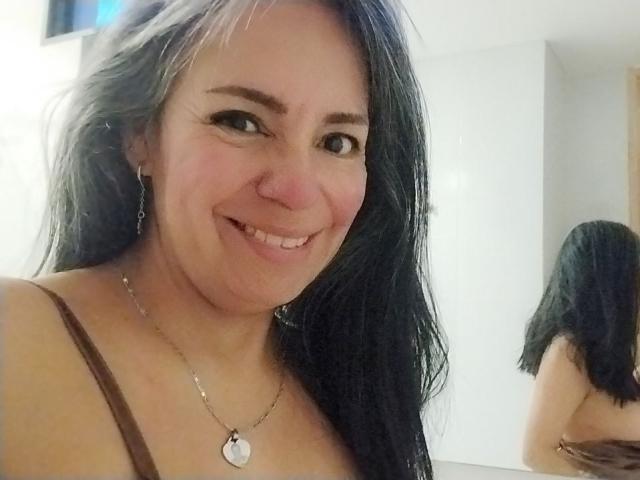 Close-up photograph of LailaCastillo with long black hair, smiling at the camera. She's wearing a brown spaghetti strap top and a silver necklace. Bright background. Her face shows visible sexual tension.
