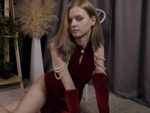 Carlotta Renzelman knielt op de vloerbedekking, gekleed in een dieprode fluwelen jurk met parelarmbanden, waarbij haar rechter dij zichtbaar is. Haar blonde haar is recht, met een grijze achtergrond. Ze poseert sexy en zelfverzekerd, met een subtiele aantrekkingskracht..