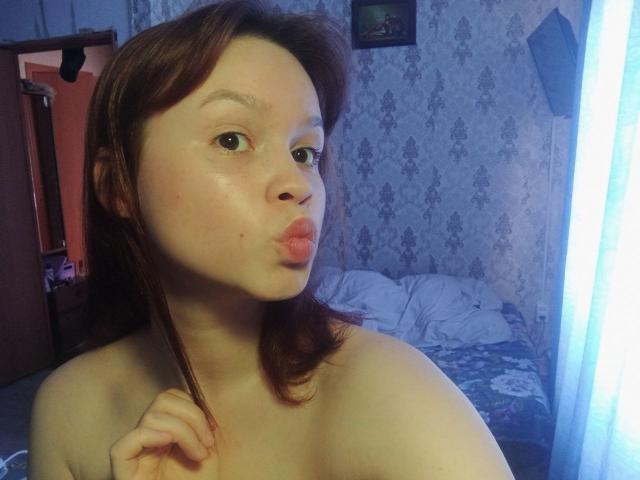 Photograph of DottieZaspel, a fair-skinned woman with brown hair, pouting her lips, topless, hand near her neck, bedroom with patterned wallpaper, unmade bed, bright light from window. Fucking sexy.