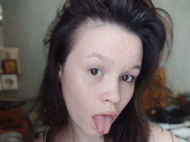 Close-up de DottieZaspel, uma mulher de pele clara e cabelo escuro, que está de língua para fora. Ela tem pequenas manchas na pele nas bochechas, cabelos levemente desalinhados e um olhar direto. Expressão muito atraente..