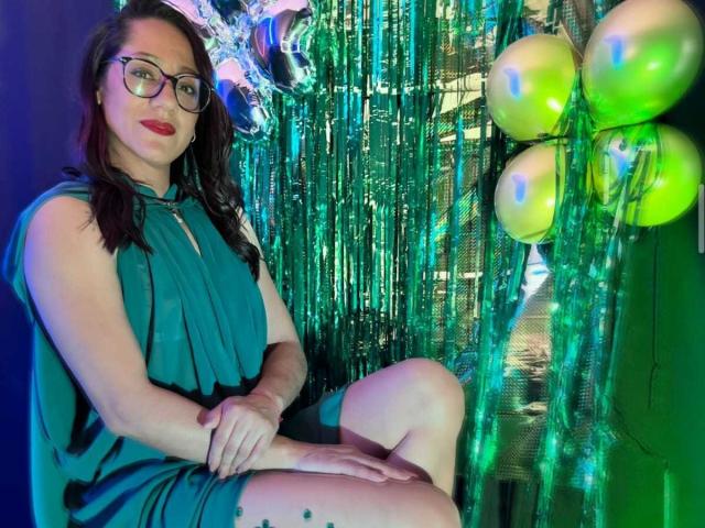 SamanthaCorine sits against shimmering green and blue metallic curtains, wearing a sleeveless teal dress, with red lipstick and glasses, legs crossed, revealing a black tattoo on her right thigh. Yellow-green balloons to the right.