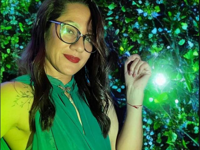 SamanthaCorine, with glasses and red lipstick, raises her right hand against a glowing green and blue leafy background. She's wearing a green sleeveless top, her dark hair flowing. Fucking hot.