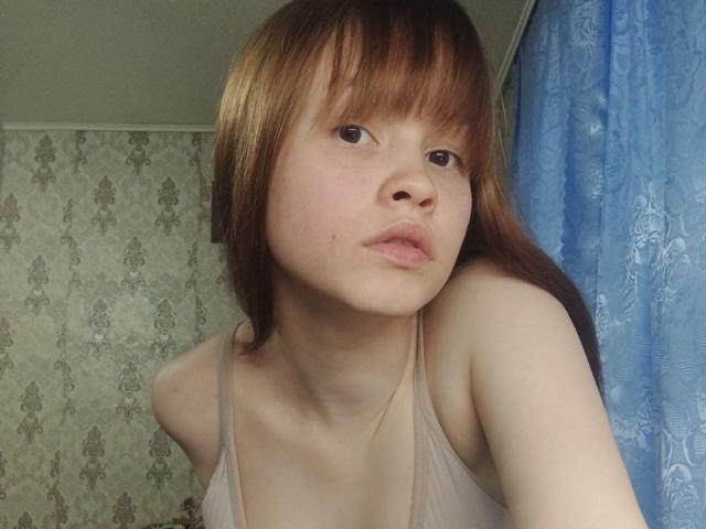 Nahaufnahme von DottieZaspel, einer jungen Frau mit heller Haut und braunen Haaren, die ein beiges Tanktop trägt und direkt in die Kamera schaut. Kamera mit leicht geöffnetem Mund. Blauer Vorhang auf der rechten Seite
.