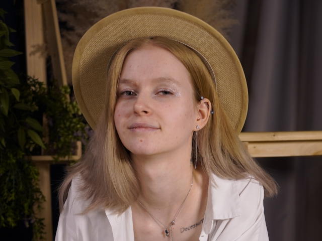 Fotografia de Carlotta Renzelman, uma jovem de cabelos castanho-claros, usando um chapéu bege e uma camisa branca, com um leve sorriso. manchas, maquiagem discreta, colar pequeno com a frase "Ouse Sonhar", reflexo de luz visível..