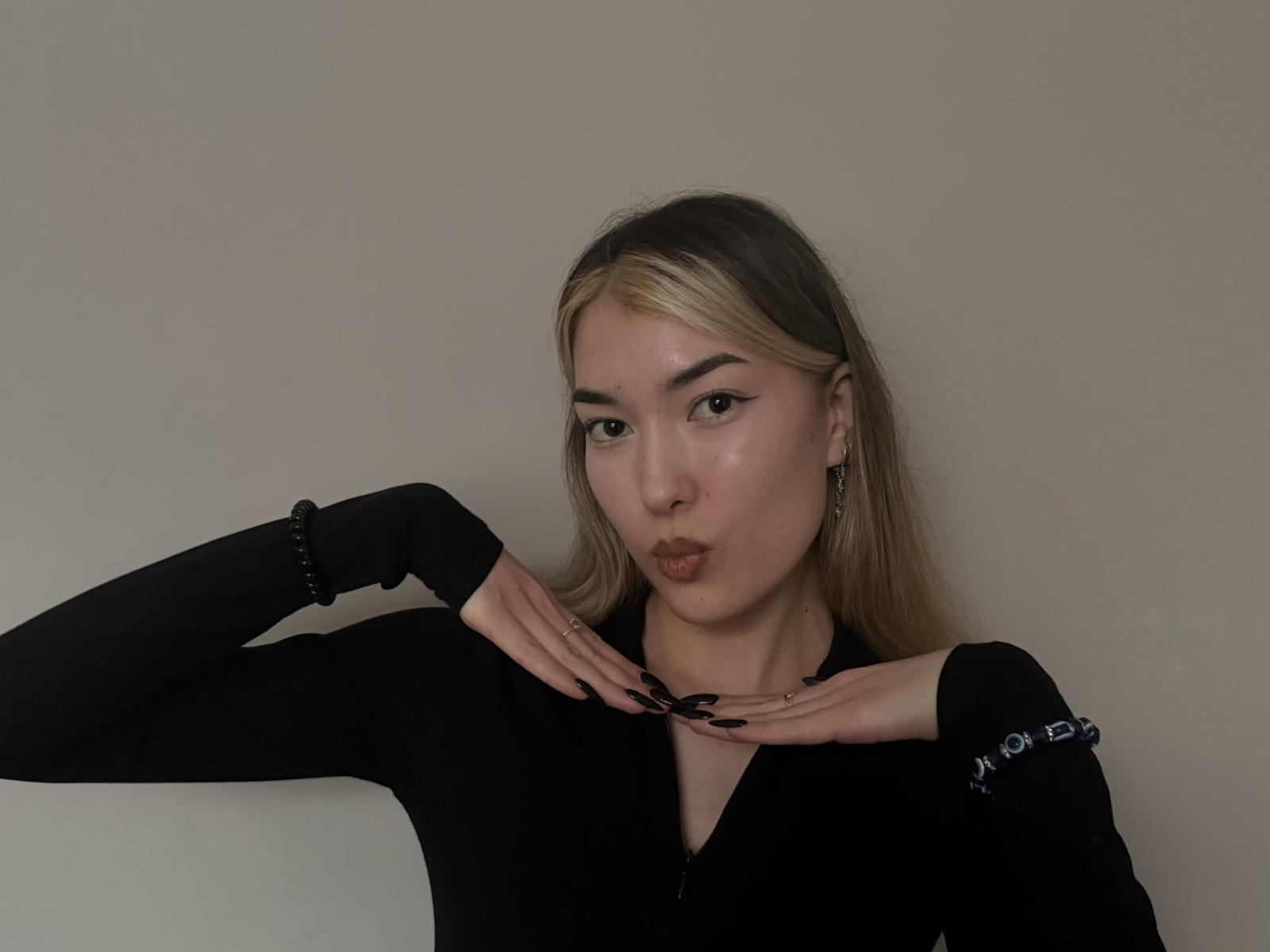 Foto van BrianneMarchese met lichtbruin haar, volle lippen en opgetrokken handen die haar nek omlijsten. Ze draagt een zwarte top met lange mouwen, kleine oorbellen. Ze heeft een lichte huid, een sensuele uitdrukking en een lichte blos. Grijze achtergrond.