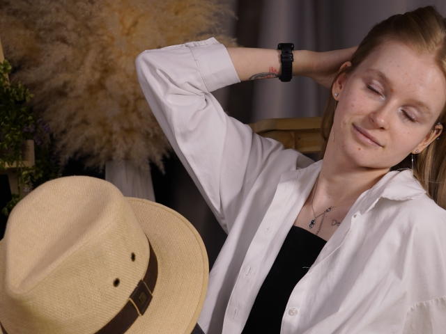 CarlottaRenzelman with closed eyes, relaxed pose, hand behind head, wearing white shirt and black top, holding beige hat with brown band, neutral expression, casual setting, soft lighting.