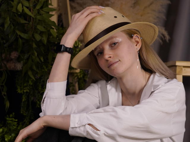 Fotografia de Carlotta Renzelman usando um chapéu de palha, camisa branca, relógio preto e brincos, apoiada em uma cadeira. mão no chapéu, sorriso confiante, iluminação fraca, folhagem verde ao fundo..