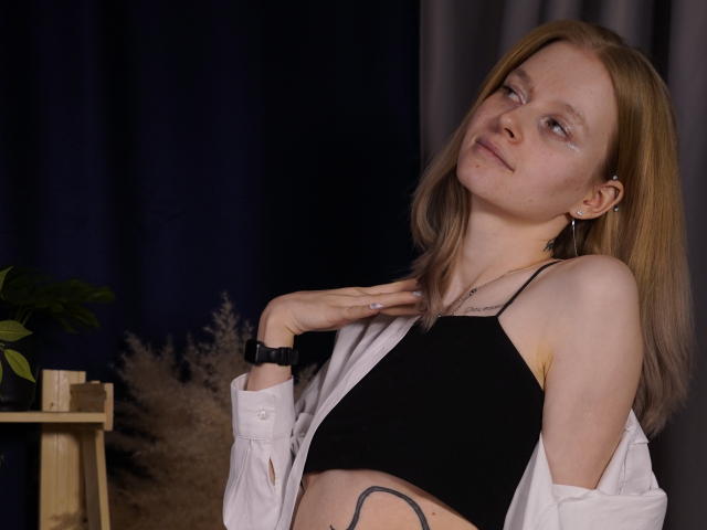 Carlotta Renzelman, met lang bruin haar, draagt een zwarte crop top en een witte, open geknoopte blouse, en kijkt naar de camera. Een lichtelijk gekanteld hoofd. Donkere achtergrond, kleine plant in de hoek. Verdomd aantrekkelijk..