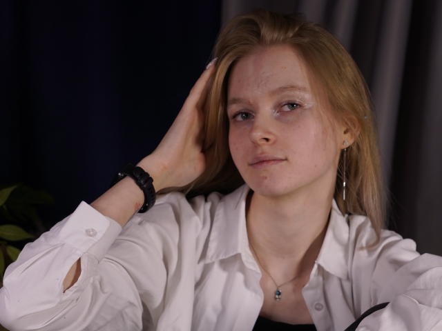 Photographie de Carlotta Renzelman, avec une peau sale, des yeux bleus et des cheveux bruns clairs, portant une chemise blanche à boutons, noire collier et pendentif en argent, regardant sur le côté, une main dans les cheveux. Extrêmement sexy..