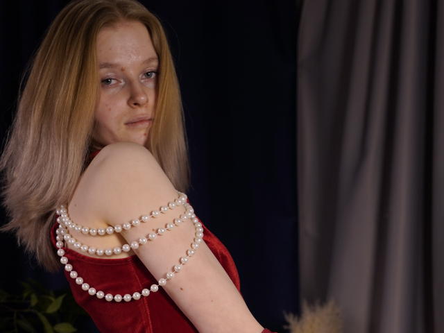 Foto van Carlotta Renzelman, met een lichte huid en sproeten, gekleed in een rode fluwelen jurk en een witte parelketting. Kijkend over haar schouder, donkere achtergrond, subtiele seksuele spanning, nonchalante pose, zelfverzekerde uitdrukking..