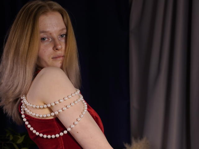 Fotografía de Carlotta Renzelman, una mujer de piel clara con cabello rubio hasta los hombros, vestida con un vestido rojo de terciopelo, drapeado. con un collar de perlas blancas, mirando por encima del hombro con una expresión seductora..