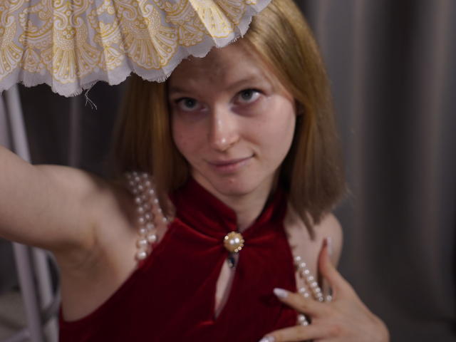 Carlotta Renzelman, com cabelo castanho curto, vestindo uma blusa vermelha com alças e detalhes de pérolas, segurando um tecido com borda de renda. Acima da cabeça dela, olhando para cima com um sorriso discreto, em uma pose sugestiva..