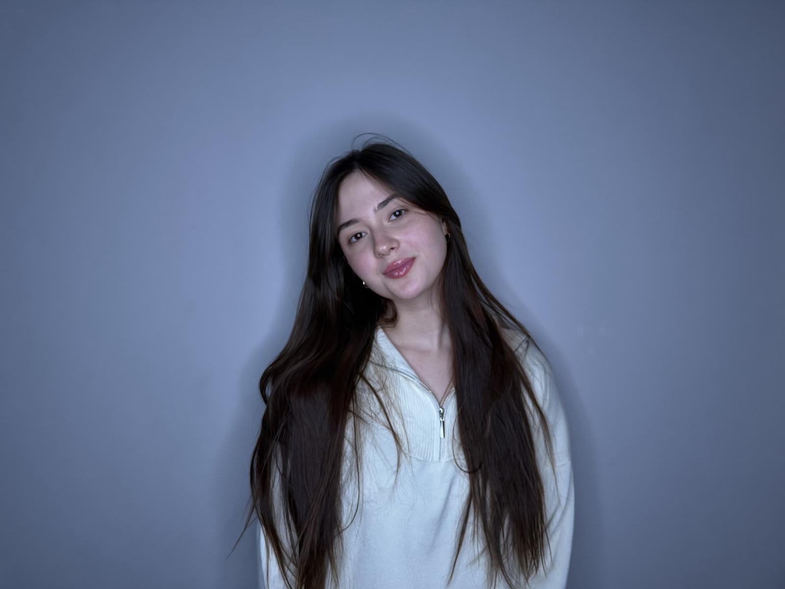 Photographie d'Emogene Busalacchi, debout devant un fond gris, avec de longs cheveux noirs et humides, portant une robe blanche chemise. Elle regarde directement l'appareil photo, la bouche légèrement ouverte, dégageant une tension sexuelle..