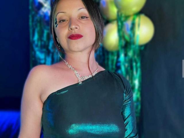 Fotografia di Angelina Blanche con capelli neri corti, rossetto rosso e un abito nero lucido con una spalla scoperta, che indossa un collana scintillante.
lo sfondo presenta palloncini gialli e verdi luminosi. Tensione sessuale, sguardo seducente..