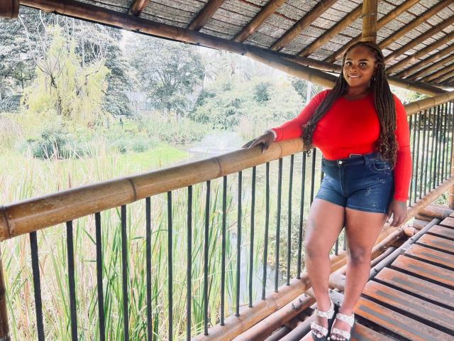 HayleyEvans, a curvy Black woman with long braids, leans against a bamboo railing, wearing a red sweater and blue denim shorts. Bright light highlights her figure, creating a sexy outdoor vibe.