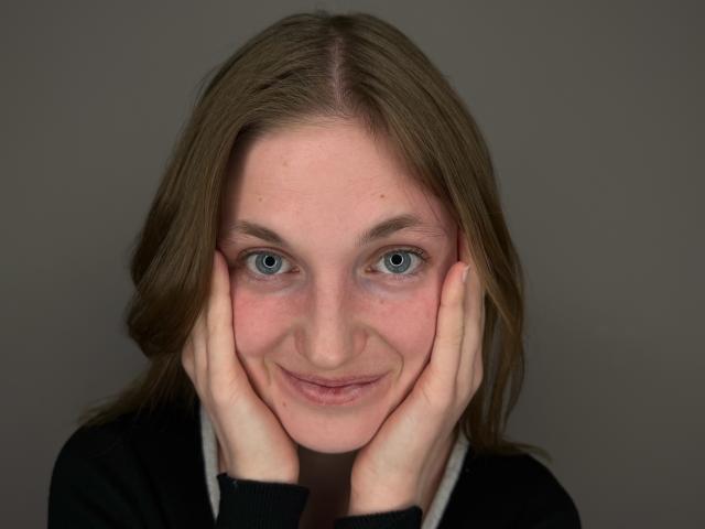ShantaeKleve a les yeux bleus, une peau claire et des cheveux bruns, elle sourit avec les mains sur ses joues, et porte une chemise noire avec un col blanc, sur une photo en gros plan..