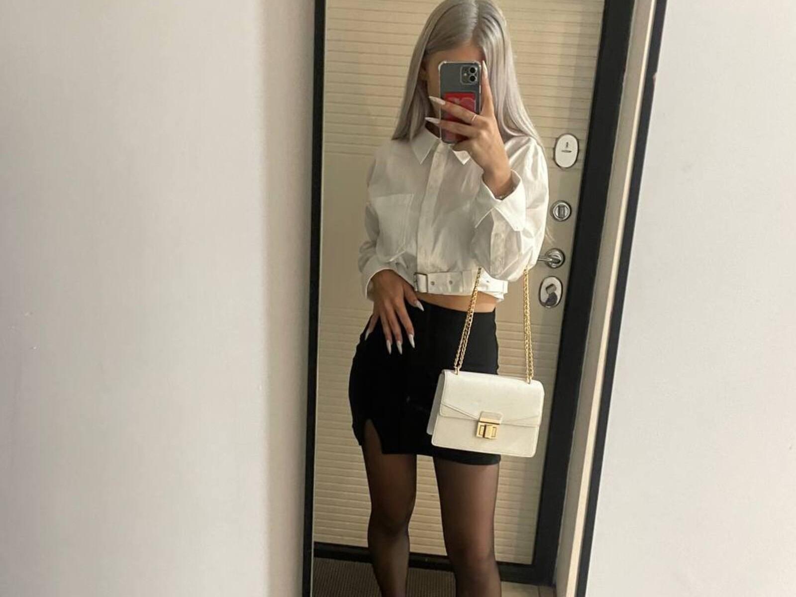 Foto van Corrinne Astrup met zilverkleurig haar, gekleed in een witte blouse en een zwarte minirok, terwijl ze iets wits vasthoudt. handtas, spiegel-selfie, telefoon voor het gezicht, zichtbare erectie..