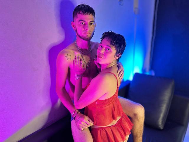 Ein interrassisches Paar sitzt auf einer schwarzen Couch, beleuchtet von violetten und blauen Lichtern. Sie trägt ein rotes Spitzenkleid, umarmt ihn von der Seite, ihre Hand liegt auf seiner Brust, sein Penis ist sichtbar..