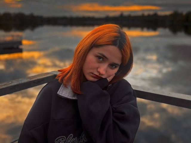 Gricelda Lydick, con cabello naranja de longitud media, apoyada en una barandilla, vistiendo una chaqueta negra con cuello blanco, un cielo al atardecer con colores dramáticos. El cabello naranja contrasta con la chaqueta oscura, evocando tensión sexual..