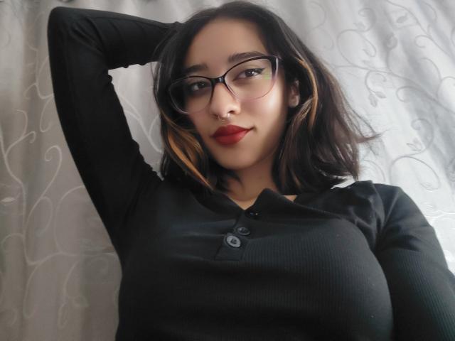 Photograph of CamiVargas with medium-length dark hair, wearing black glasses and a tight black shirt highlighting her large breasts. She raises her left arm, with a seductive look and red lipstick.