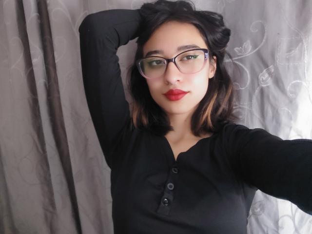 Photograph of CamiVargas with shoulder-length black hair, red lipstick, and black button-up shirt, wearing glasses, hand behind head, neutral gray curtain background, slight light shadow on right side.