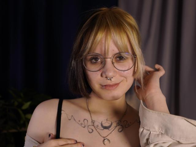 VickeyParkes tem cabelo castanho, na altura dos ombros, com mechas loiras, usa óculos redondos e uma blusa preta com alças. Ela tem tatuagens no peito e um piercing no nariz. A mão dela toca o rosto, mostrando um leve sorriso..