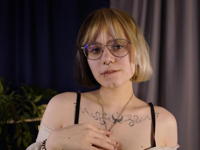 VickeyParkes has a short blonde bob, wears glasses, and has a nose ring. She's topless, showing black bra straps, a chest tattoo, and holding a necklace. Fucking hot, with a serious look.
