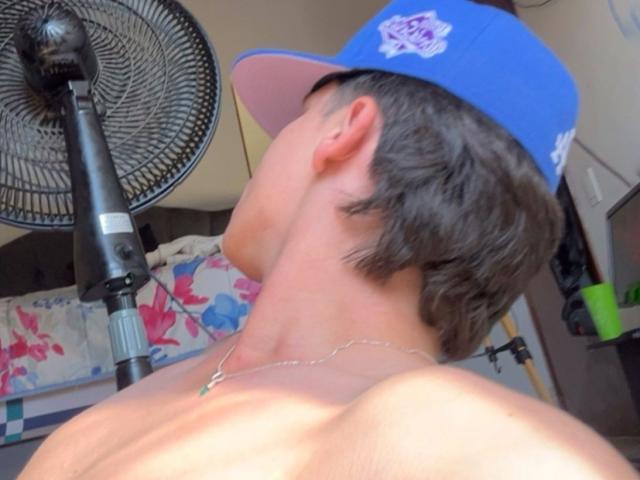 SebastiaQuesada senza camicia, con capelli castani corti, un berretto blu e una collana, si sporge in avanti, con la bocca aperta, vicino a una ventola nera. La pelle brilla, tesa, con un'atmosfera carica di sensualità. Colori vivaci sullo sfondo..