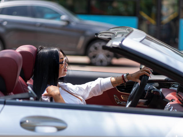 BlackClover with long black hair, wearing a white off-shoulder top, sits in a red convertible, gripping the steering wheel, looking forward with sunglasses on, intense sexual tension in the air.