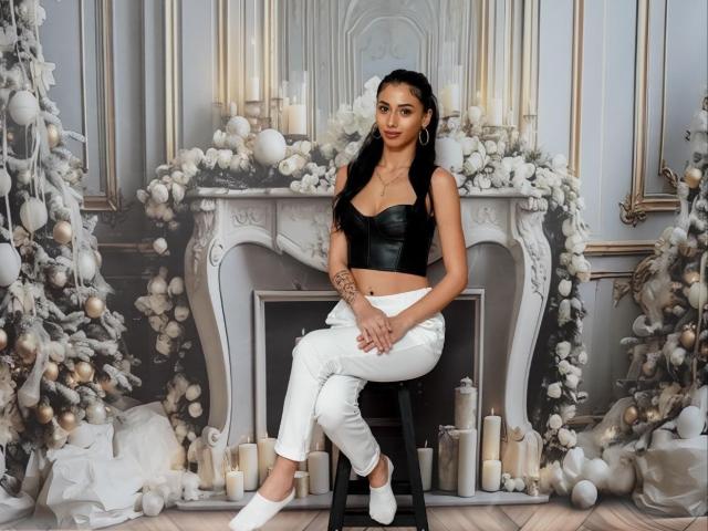 Tia Cyrus, zittend op een kruk, draagt een zwart leren topje en witte broek, met lang zwart haar, sexy, wit. Kerstversieringen en kaarsen op de achtergrond.