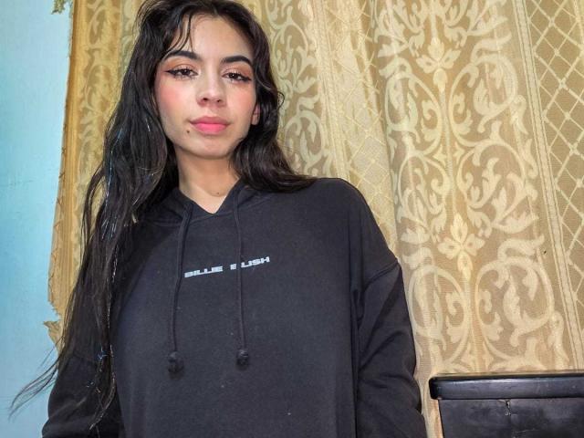 Photograph of KatherinJohns, standing in front of ornate gold curtains. She has long black hair, pink lipstick, and is wearing a black hoodie with "JUICE" in white text. Fucking hot, cock-teasing look.