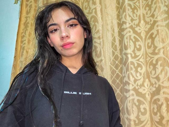 Photograph of KatherinJohns, with long black hair, wearing a black "Bale & Luxe" hoodie, standing against a gold damask curtain. Her red lipstick contrasts with her pale skin, creating a sexy look.