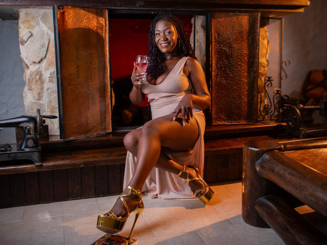 DianethKiss está sentada con confianza en una habitación con poca luz, vestida con un vestido blanco de un solo hombro y tacones de plataforma dorados. Ella sostiene una copa de vino tinto, con las piernas cruzadas de forma seductora. Una cálida luz anaranjada..