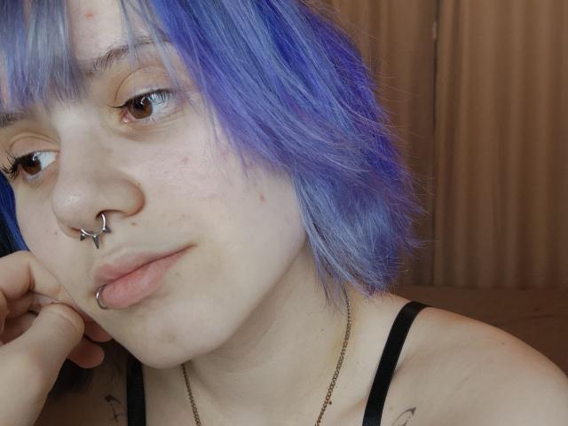 Close-up foto van VickeyParkes met blauw geverfd haar, een neusring en een lipring, met haar kin op haar hand rustend, gekleed in een zwarte bandeau. Boven. De huid is bleek met lichte sproeten, kijkt weg..