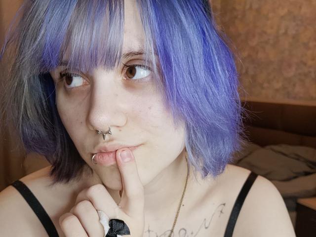 Foto em close-up de VickeyParkes com cabelo curto azul-roxo, olhos castanhos, piercing no nariz e piercing no lábio. VickeyParkes morde o dedo de forma sedutora, usando tiras pretas, com tatuagens no peito e pele clara. Tensão sexual.