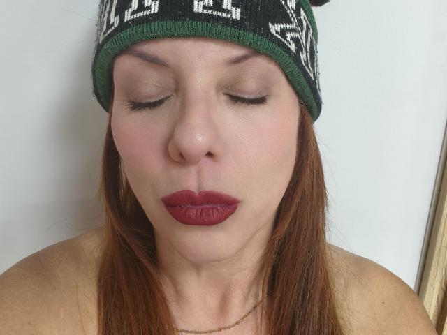 Fotografia ravvicinata di Khharha, una donna bianca con capelli rossi, che indossa un berretto nero con scritte bianche, con gli occhi chiusi, e con un'espressione seria. rossetto, spalle scoperte, labbra leggermente dischiuse, tensione sessuale, sfondo neutro.