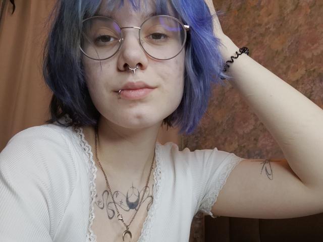 VickeyParkes tiene el pelo azul, gafas redondas y un piercing en la nariz. Ella lleva una blusa blanca con encaje, que deja ver tatuajes en su pecho y brazo. Ella está posando con la mano en el pelo, mirando directamente a la cámara..