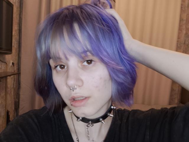 VickeyParkes tem cabelos azuis e roxos desgrenhados, um piercing no nariz e um piercing no lábio. Usando um colar preto com espinhos, segurando o cabelo com uma mão. Pele clara, olhar intenso.