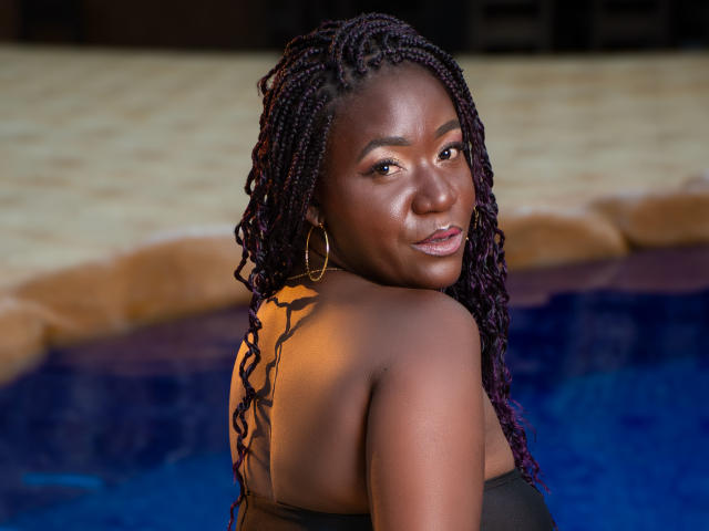 Photograph of DianethKiss, dark-skinned woman with long, braided hair, looking over her shoulder, glowing skin, hoop earrings, topless, blue pool in background, intense gaze, slight smile, wet hair, sexual tension.